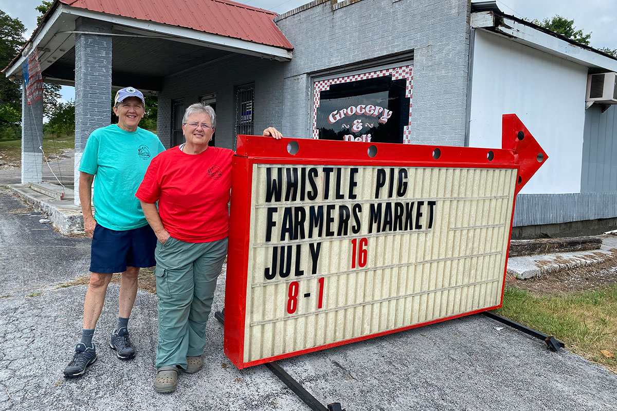 Jan Brown and Melissa Hubbard standing next to a sign that says "Whistle Pig Farmers Market" in front of the old cement block Grocery & Deli store.