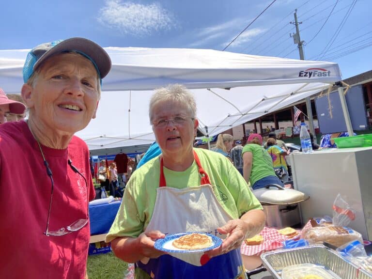 Jan Brown and Melissa Hubbard serving pancakes outside a tent on a sunny day.